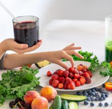 A glass of smoothie from berries in a child's hand on the background of a kitchen table with fruits and vegetables, making a smoothie at home.